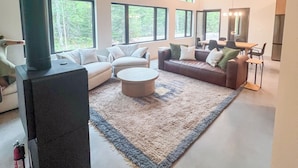 A spacious living room with a large black fireplace, a ceiling fan, and multiple windows offering a view of the outdoors. - Spruce House: The Perfect Summer Retreat with Hiking, Swimming, and More (Copper Harbor)