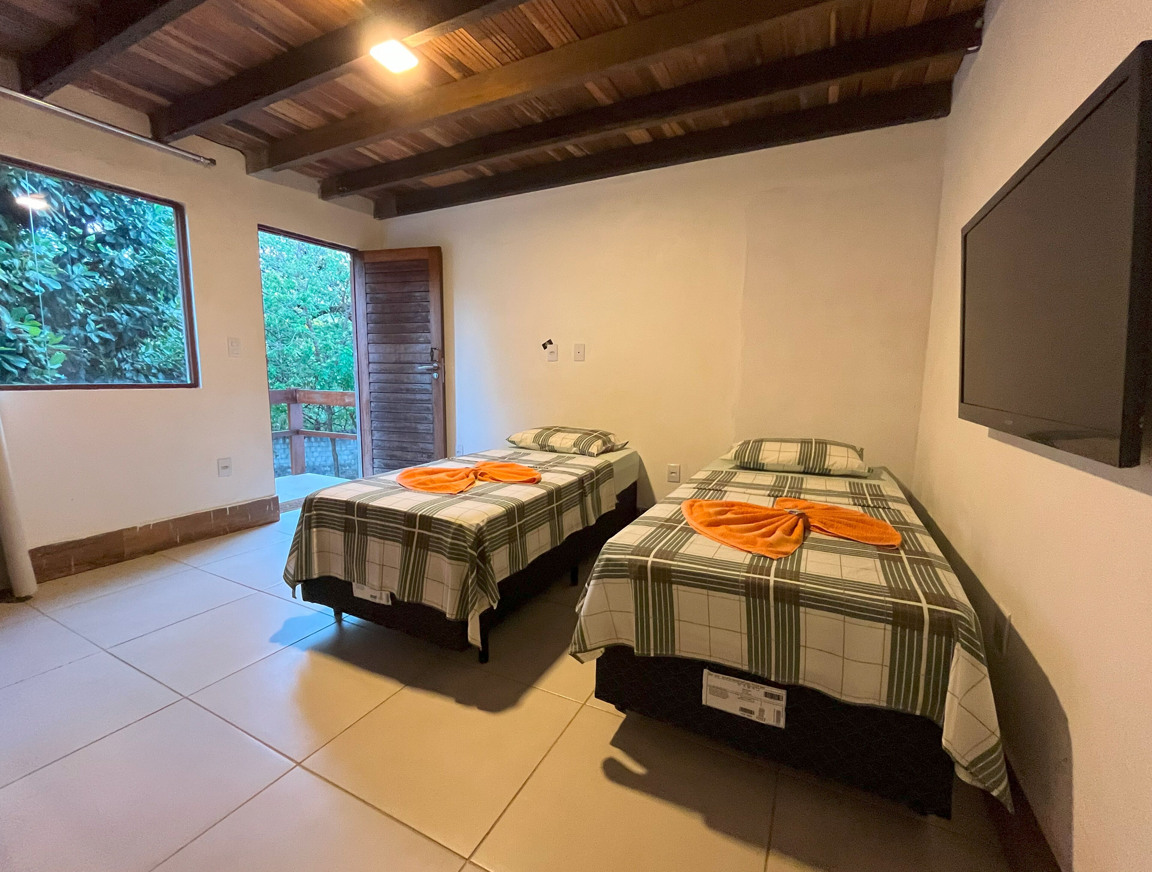 Quarto com duas camas, uma janela grande, teto de madeira e televisão na parede.