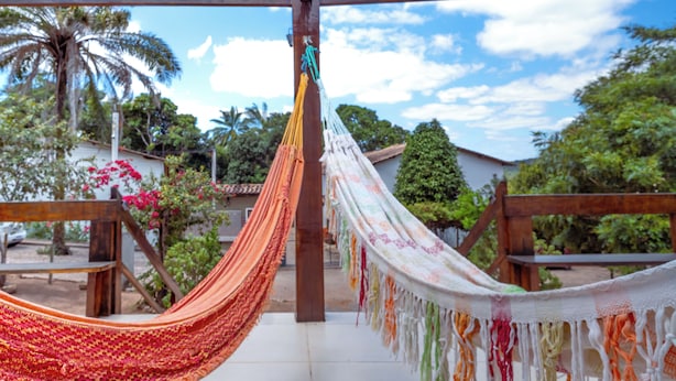 A hammock suspended between wooden beams in a garden setting with palm trees and a house in the background. - Pousada em Lençóis Reserva do Dendê (Lençóis)