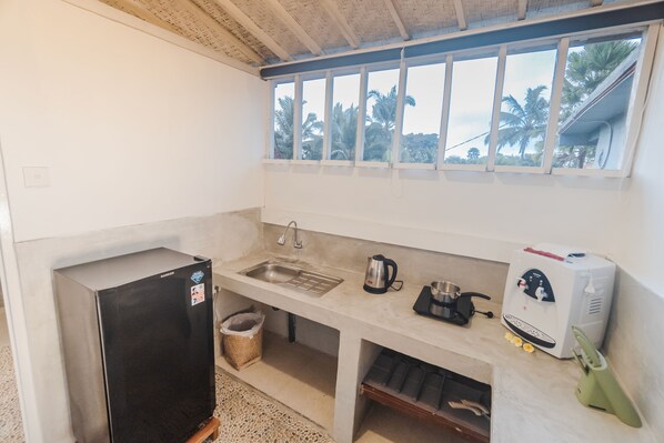A compact kitchen with a refrigerator, sink, kettle, coffee maker, and a water dispenser.