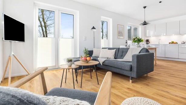 A modern living room with a grey sofa, a coffee table, and a TV on a tripod. There is a kitchen area with white cabinets and a dining table.