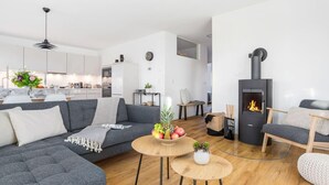 A modern living room with a grey sofa, a small wooden table with fruit, a black wood-burning stove, and a kitchen in the background.