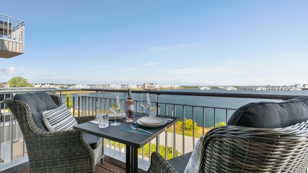 A balcony with wicker furniture and a table set for two, overlooking a body of water and a cityscape. - Stunning apartment in Olpenitz (Olpenitz)
