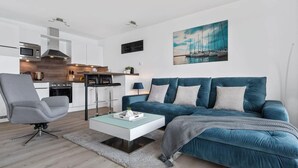 A modern living room with a blue sofa, a grey armchair, and a glass coffee table. A kitchen with white cabinets and a microwave is visible in the background. - Stunning apartment in Olpenitz (Olpenitz)