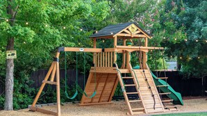 Une structure de jeu en bois comprenant un toboggan, un filet d’escalade et un petit abri.