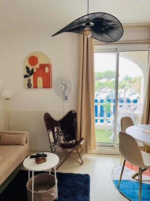 Ein Wohnzimmer mit einer Couch, einem Sessel, einem kleinen Tisch, einem Ventilator und Blick ins Freie.