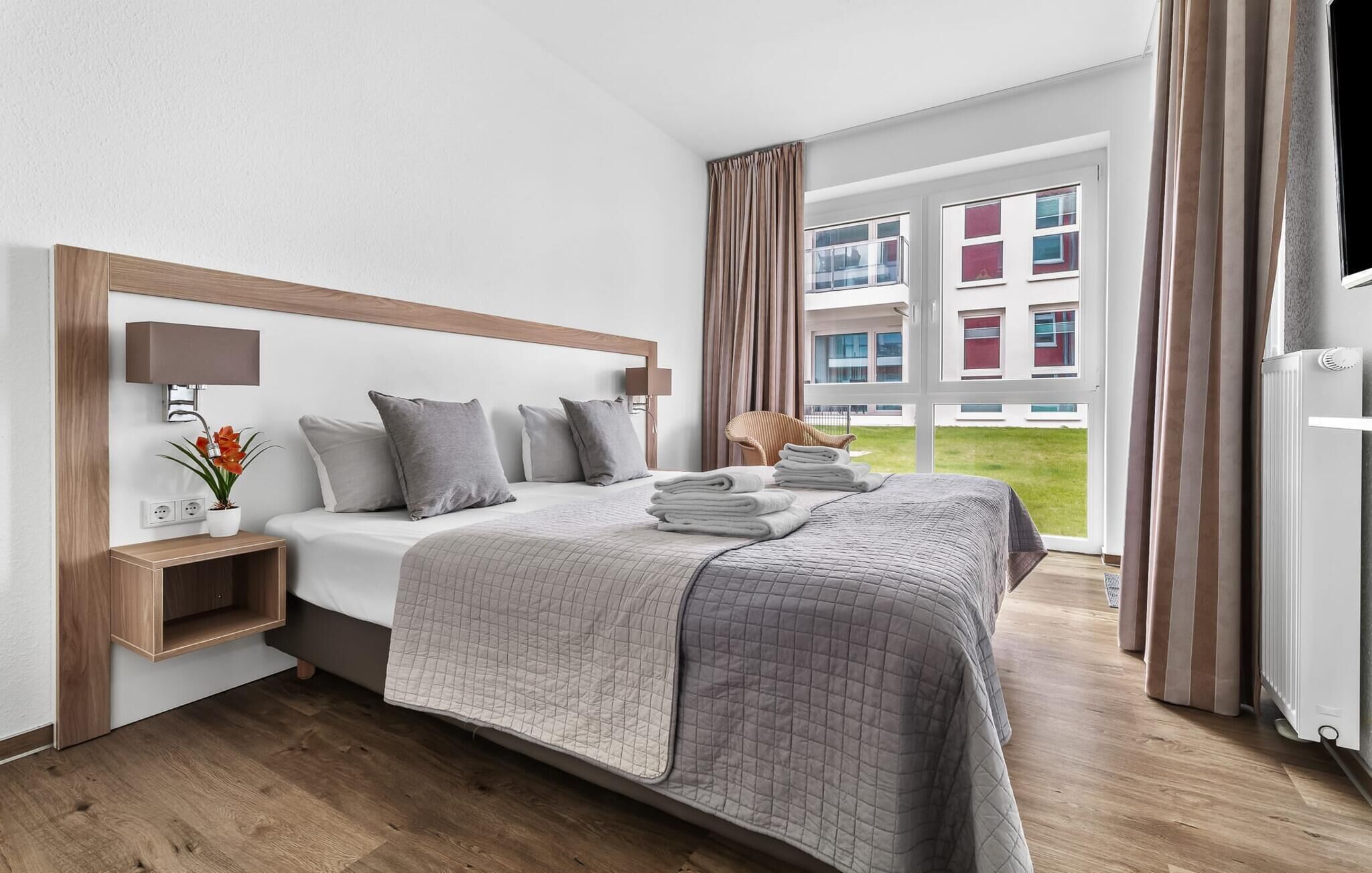 A hotel room with a large bed, a bedside table with a lamp, a chair, and a view of a building through the window.
