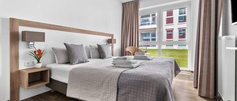 A hotel room with a large bed, a bedside table with a lamp, a chair, and a view of a building through the window.