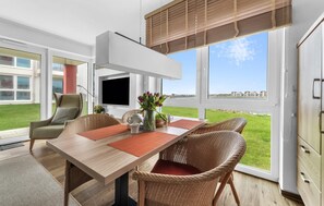 A modern dining area with a wooden table, wicker chairs, and a large window offering a view of the sea and buildings. - Gorgeous apartment in Olpenitz with sauna (Olpenitz)