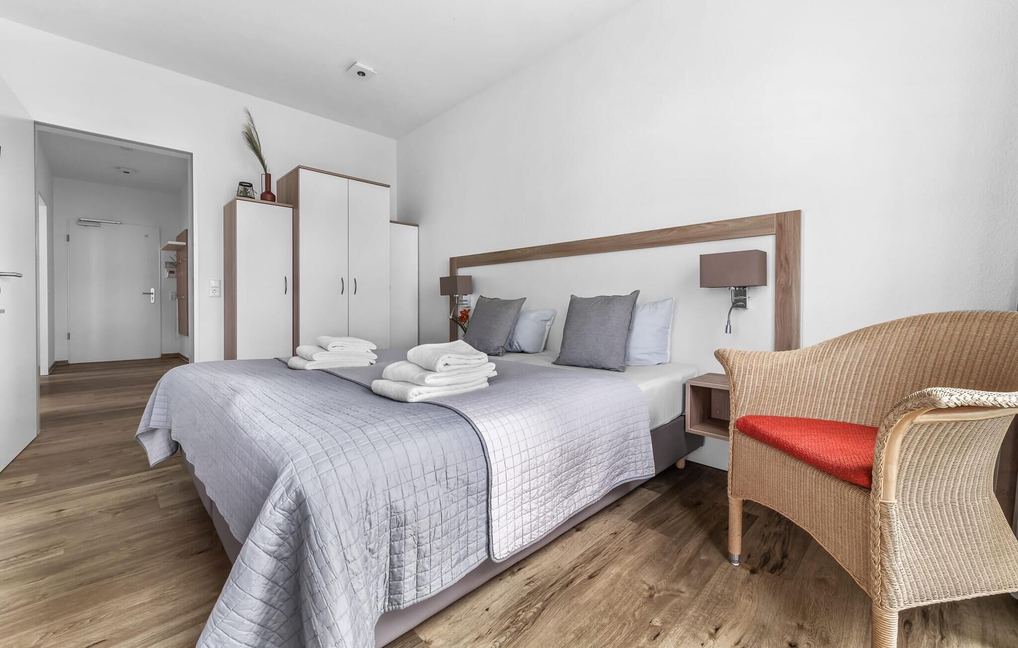 Une chambre d’hôtel moderne avec un grand lit, une armoire en bois, un fauteuil en osier et une table de chevet avec une lampe.