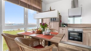 Ett modernt kök med utsikt över havet, en matplats med flätade stolar och ett träbord med en röd bordsduk.