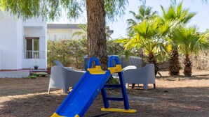 Un parque infantil con un tobogán azul y amarillo, un árbol y un edificio blanco al fondo.