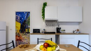 Una mesa de comedor con un frutero, un cuadro en la pared, una zona de cocina con electrodomésticos y un armario blanco.