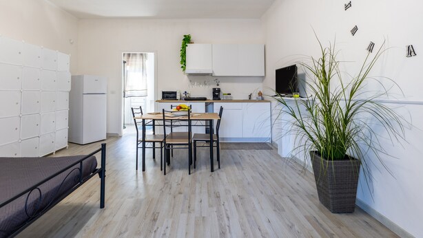 Una cocina compacta con armarios blancos, una mesa de comedor con sillas, una cama con cabecera y una planta en maceta.
