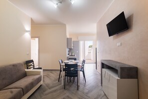 A living room with a sofa, a dining table with chairs, a flat-screen TV mounted on the wall, and a kitchen area in the background. - Apartment 'i Portici Unità 101' with Balcony and Wi-Fi (san pietro in bevagna)