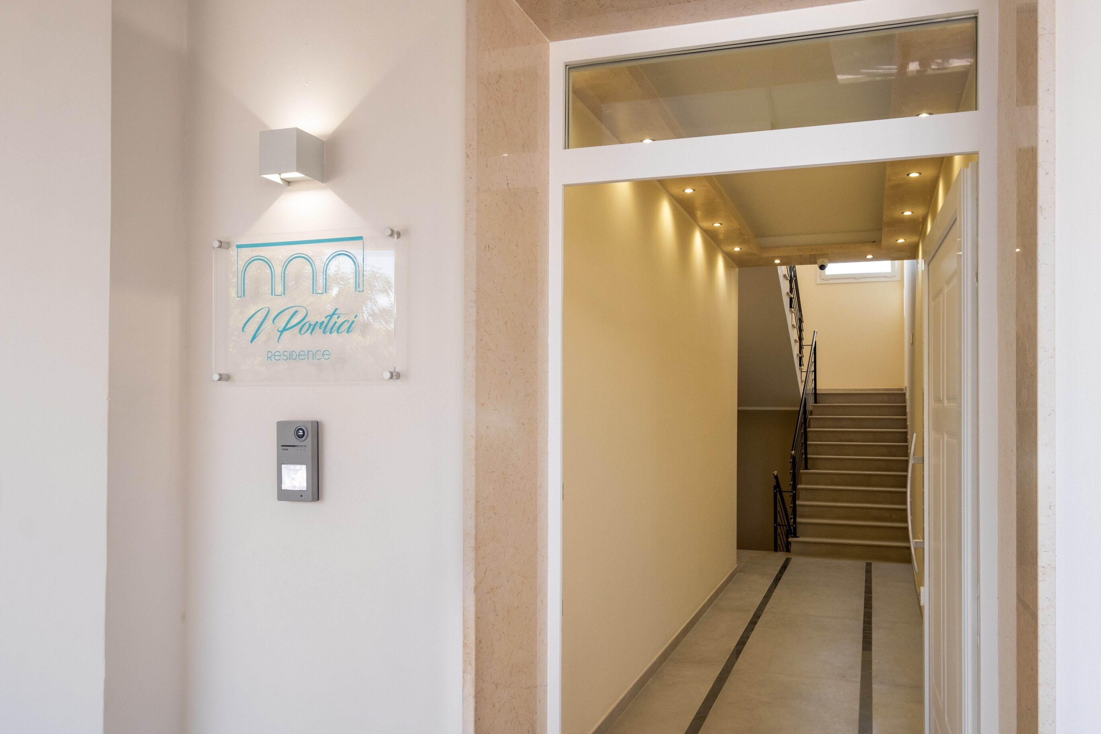 Ein Flur mit einem Schild, das 'V Portici Residence' zeigt, einem Sicherheitspanel und einer Treppe, die zu einer oberen Etage führt.