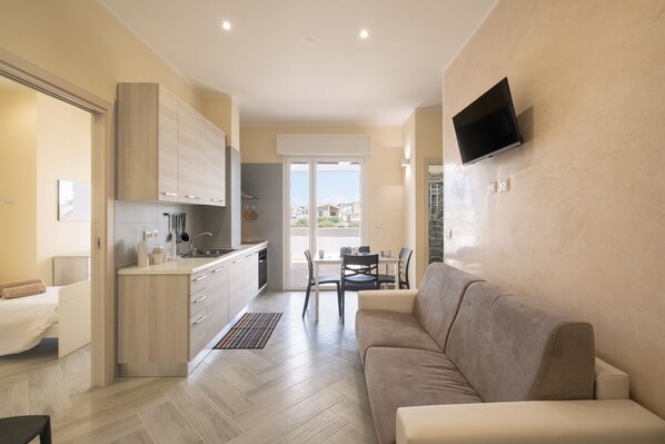 A modern living room with a sofa, a TV mounted on the wall, and a kitchen area with cabinets and a dining table.