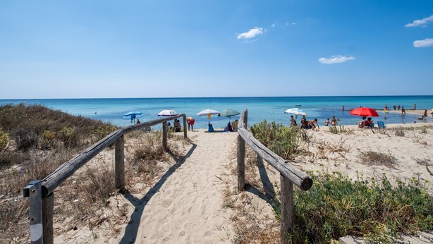 Un sendero en la playa que conduce al mar, con barandillas de madera y sombrillas.
