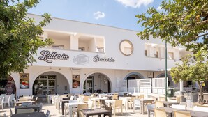 Un área de café al aire libre con mesas y sillas, un edificio blanco con arcos y señalización para 'Latteria Stella Braceria'.