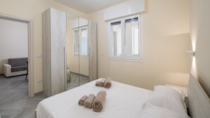 Habitación de hotel con cama, mesita de noche, espejo, ventana y puerta que da a otra habitación.