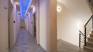 Un pasillo de diseño moderno, con piso y paredes en tonos claros, techo iluminado en azul y escalera con barandilla negra.