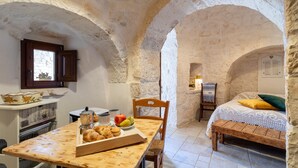 A rustic stone room with a wooden table, a bed with a green pillow, and a stone wall.