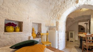 A rustic stone-walled room with a wooden table and chairs, a woven bench, and a shelf with yellow vases.