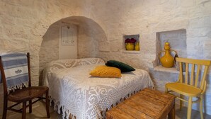 A room with a stone wall, a bed with a white lace bedspread, a wooden chair, a wooden table, and a yellow vase on a shelf.