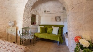 Una habitación con pared de piedra, un sofá verde, una silla de madera, una mesita de noche y una cama con colcha estampada.