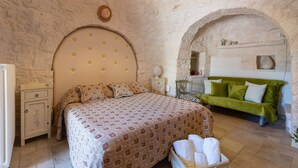 Un dormitorio con pared de piedra, cama con ropa de cama estampada, sofá verde y mesita de noche con lámpara.