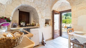 A stone-walled kitchen with a dining area, a small island, and a view through an open door. - Null 'Il Fienile' with Shared Terrace, Wi-Fi and Air Conditioning (Locorotondo)