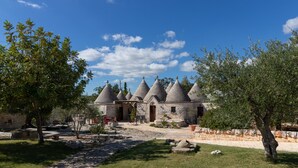 A stone building with multiple conical roofs, a paved courtyard, and surrounding greenery. - Null 'Il Fienile' with Shared Terrace, Wi-Fi and Air Conditioning (Locorotondo)