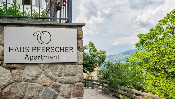 Schild für 'HAUS PFERSCHER Apartment' mit einer Wand aus Stein im Hintergrund und Blick auf Grünflächen und Berge.