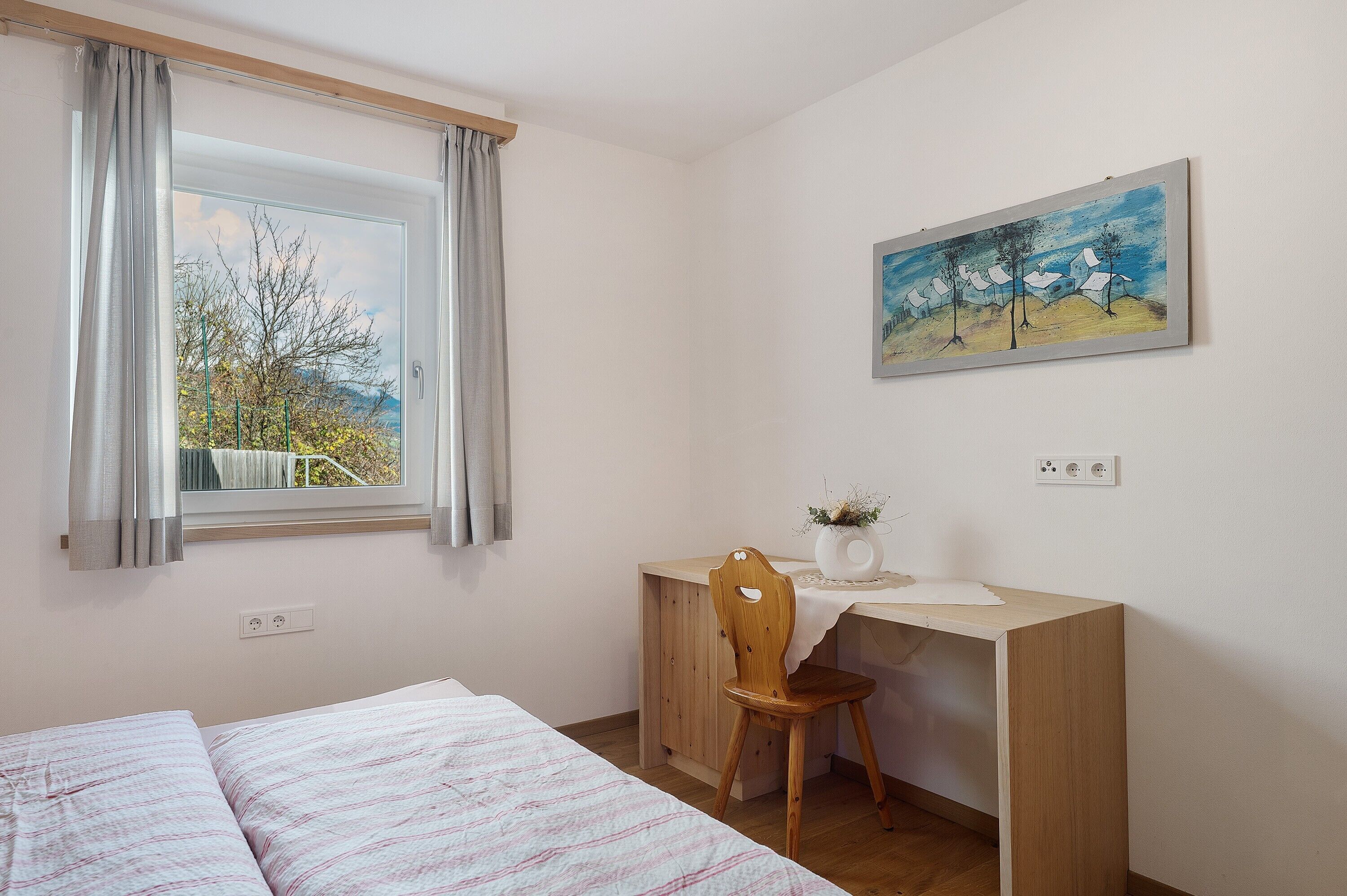 A bedroom with a bed, a desk, a chair, a window with curtains, and a painting on the wall.