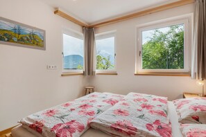 A bedroom with two beds, a window with curtains, a small stool, and a painting on the wall.