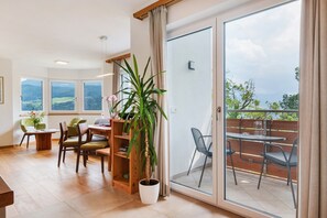 A room with a wooden floor, a dining area with chairs and a table, a potted plant, and a sliding glass door leading to an outdoor balcony with a view of the landscape.