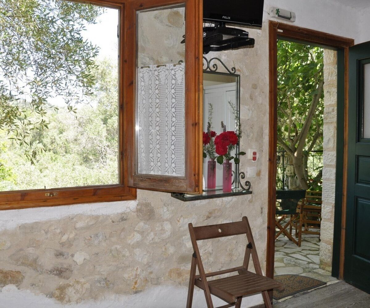 Une pièce rustique avec une chaise en bois, une fenêtre donnant sur un paysage verdoyant, un miroir agrémenté d’un rideau à motifs et une porte menant à l’extérieur.