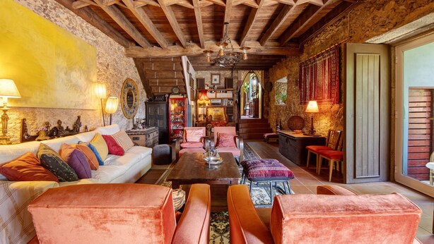 A rustic interior with exposed wooden beams, a sofa with colorful cushions, a coffee table, and a dining area with chairs.