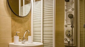 A bathroom with a sink, mirror, toilet paper dispenser, shower, and a control panel for the shower.