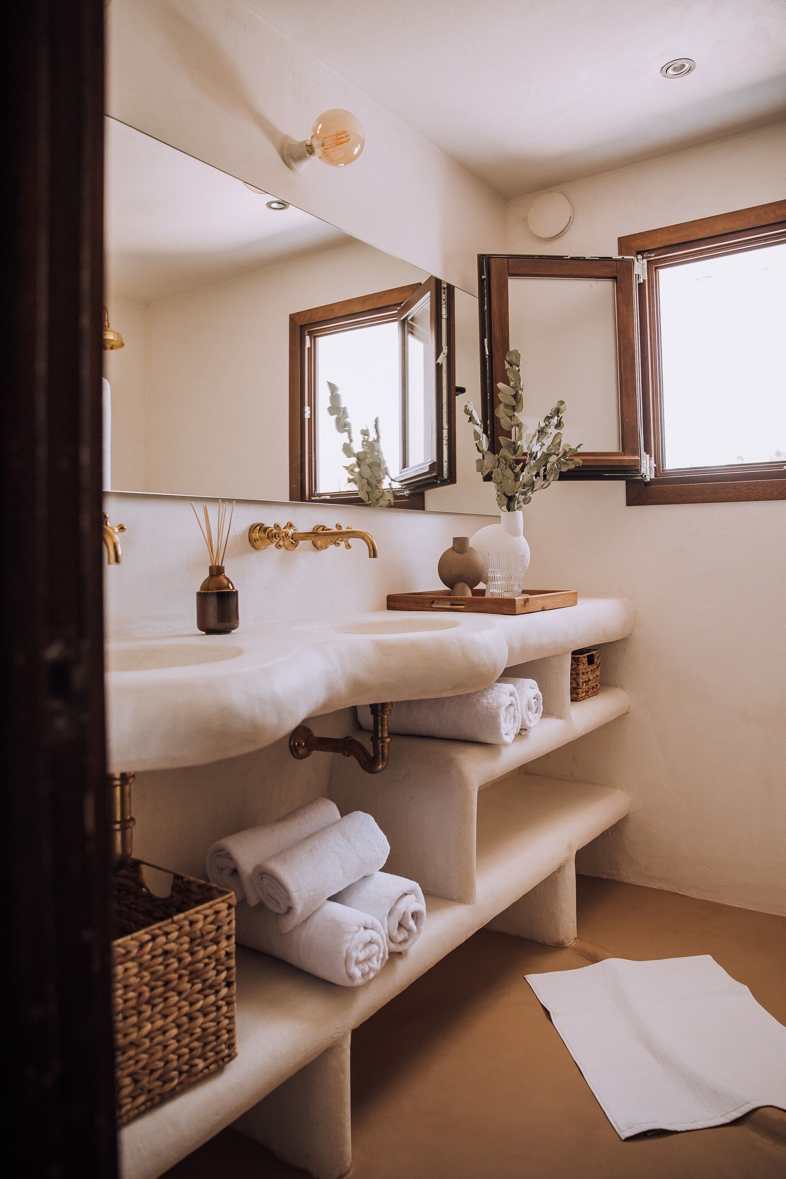 A bathroom with a white sink, two mirrors, a vase with greenery, and towels on shelves.