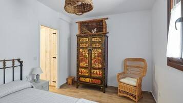 Un dormitori amb un llit, una cadira de vimet, un armari amb obres d'art i una làmpada penjant.