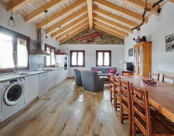 A spacious kitchen with a high ceiling, wooden beams, a washing machine, and a dining area with a table and chairs.