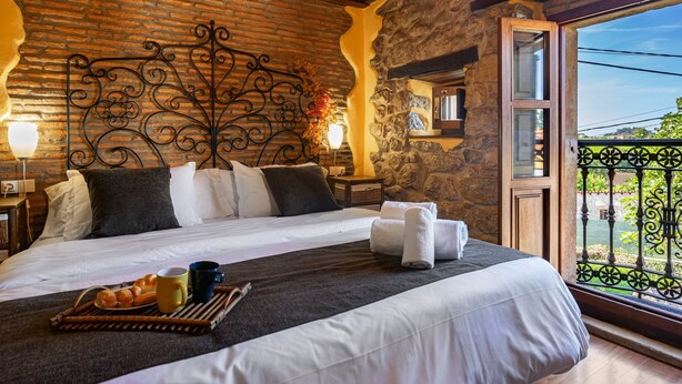 A bedroom with a stone wall, a wrought iron headboard, a bed with white linens, a balcony with a wrought iron railing, and a breakfast tray with croissants and a mug. - Country House 'El Llosín' with Private Terrace, Balcony and Wi-Fi (Ribadesella)