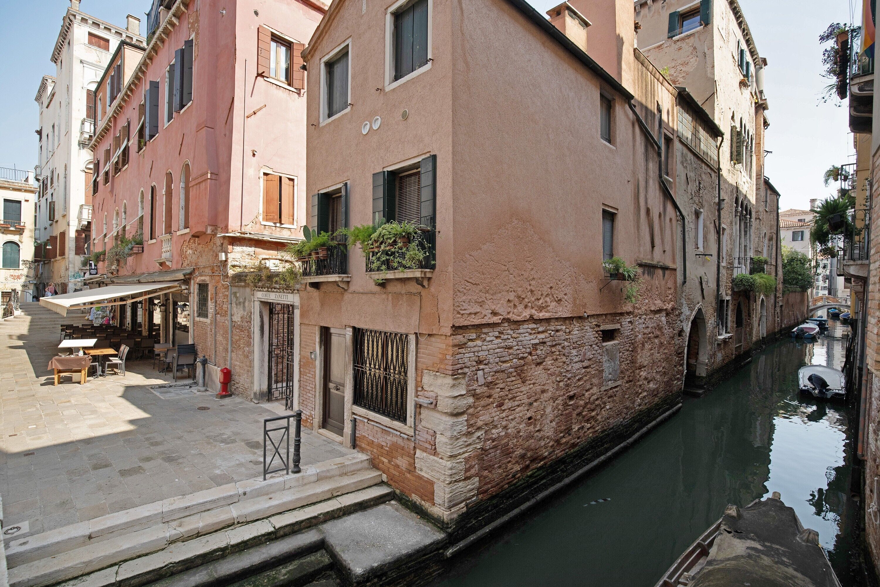 Un canal étroit, bordé de bâtiments des deux côtés, un petit espace extérieur avec des tables et des chaises, et un bateau amarré sur la droite.