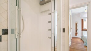 Ein Flur mit Duschkabine aus Glas, Holzboden und einem Bett im Hintergrund mit gemusterten Betttextilien.