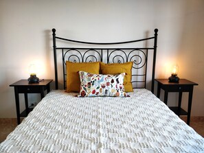 A bed with a white textured bedspread, two mustard-colored pillows, and a decorative pillow with floral patterns. There are two bedside tables with lamps on either side of the bed.