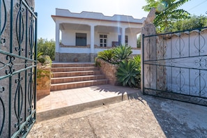 A house with a stone staircase leading to a white door, surrounded by greenery and a metal gate.
