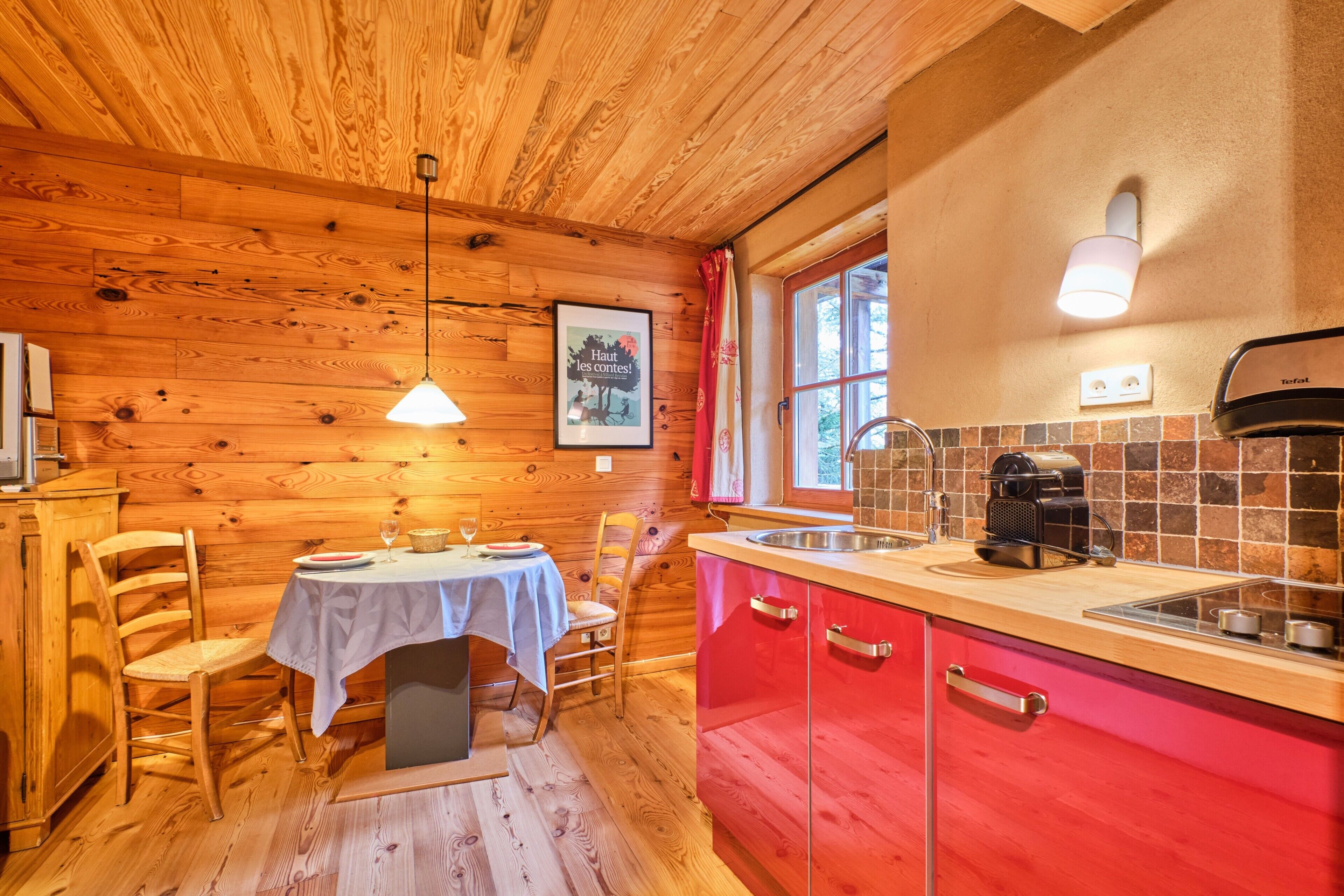Un intérieur en bois comprenant une table à manger, des chaises, une cuisine avec des placards rouges, une machine à café et une fenêtre avec des rideaux.