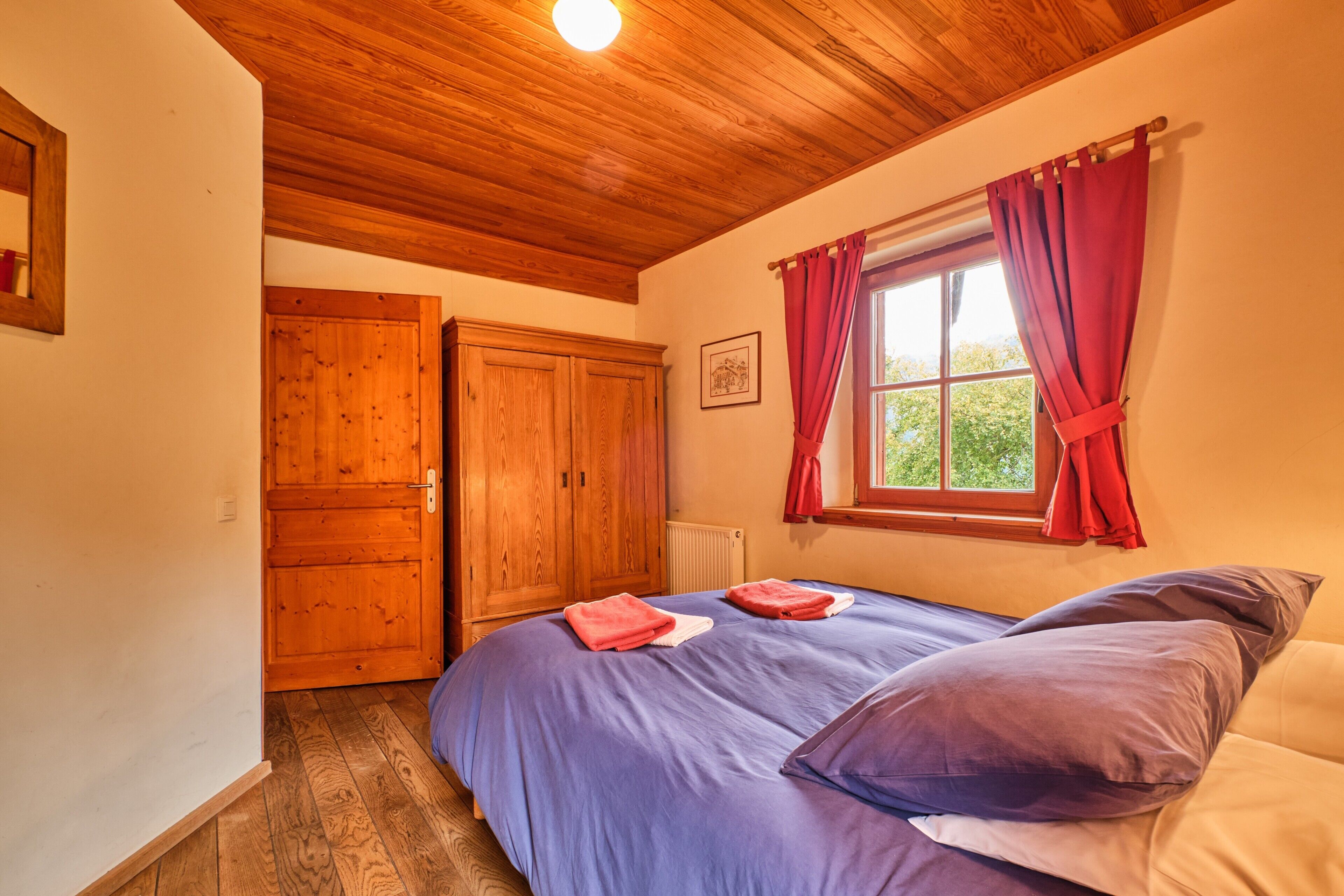A bedroom with a wooden ceiling, a window with red curtains, a bed with blue bedding, and a wooden wardrobe.