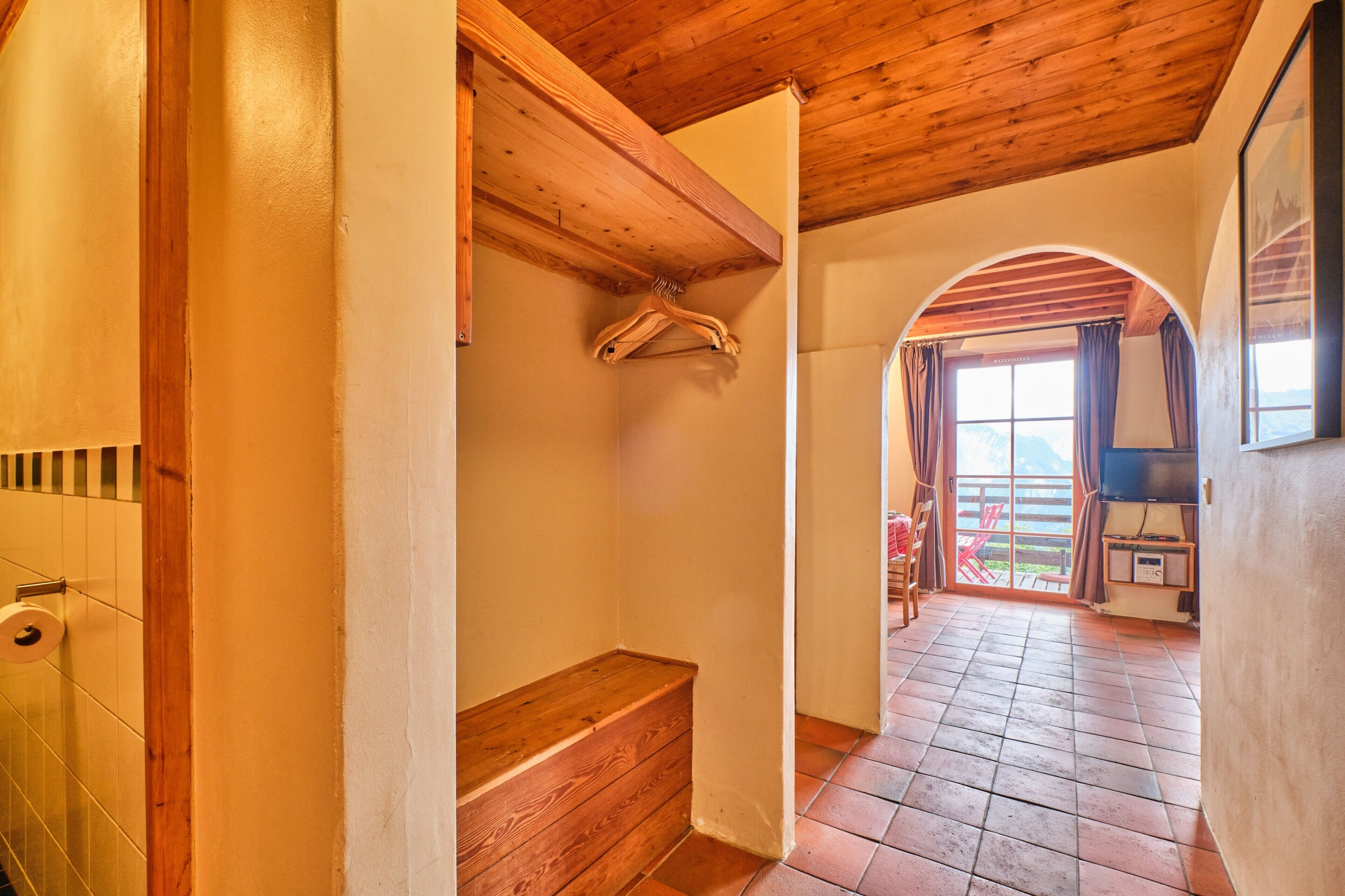 A hallway with wooden beams, a bench, a hanging coat rack, and a view through an arched doorway to an outdoor area with a TV and a red chair.
